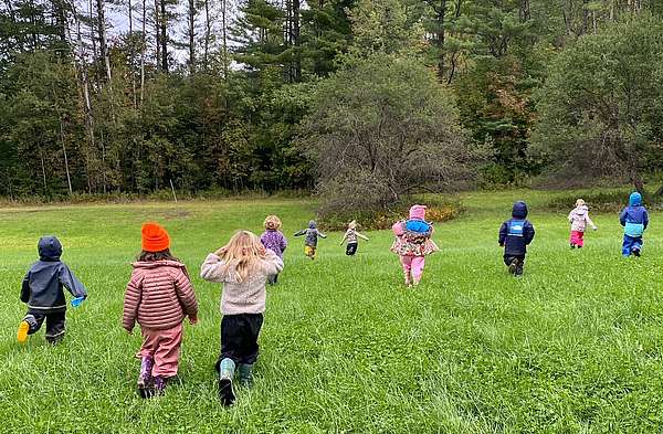 Backs to the camera, a group of kindergarteners run and disperse to explore a grassy field on the edge of a forest.