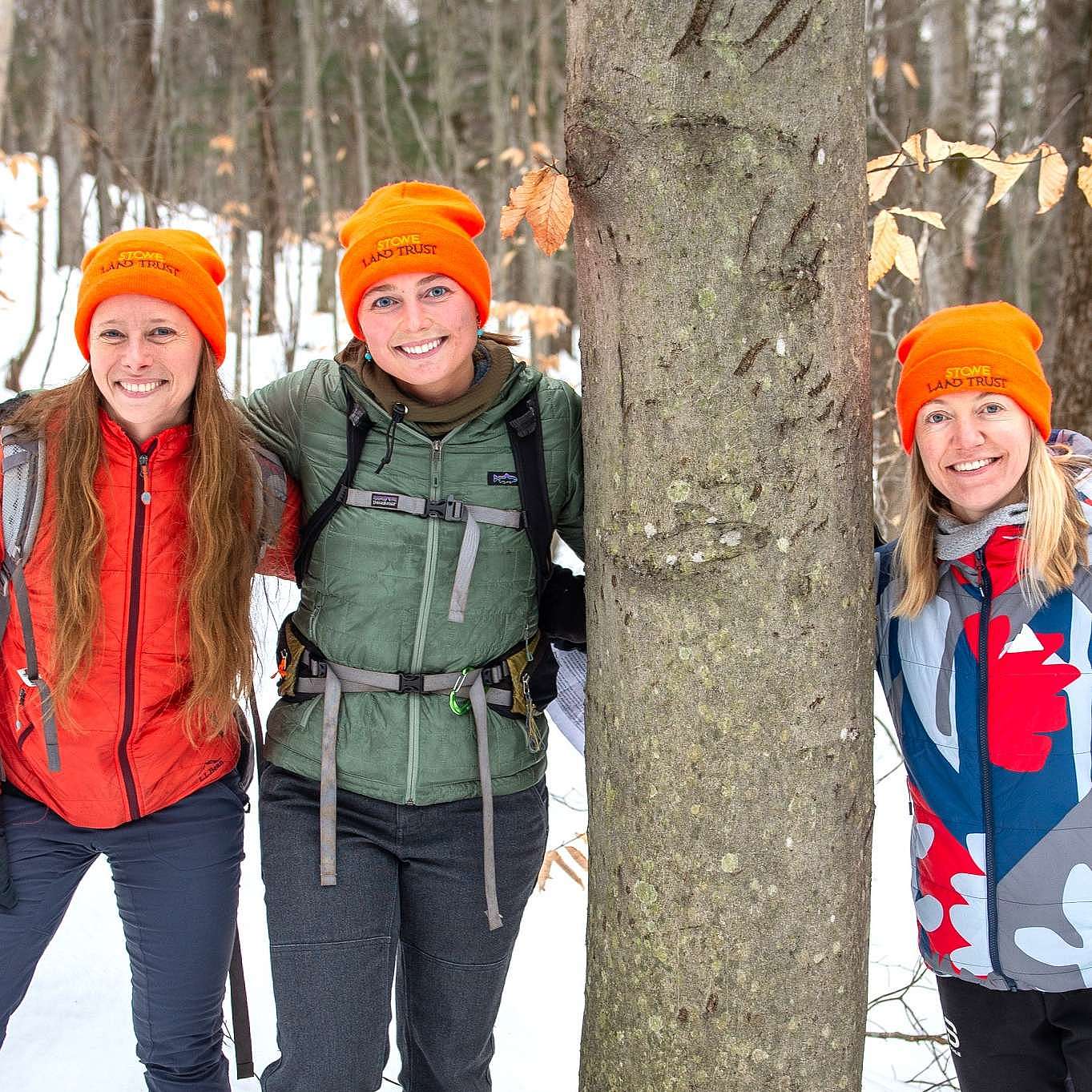 Three members of the Stowe Land Trust team, all wearing blaze orange beanies, smile next to a bear-scarred beech tree.